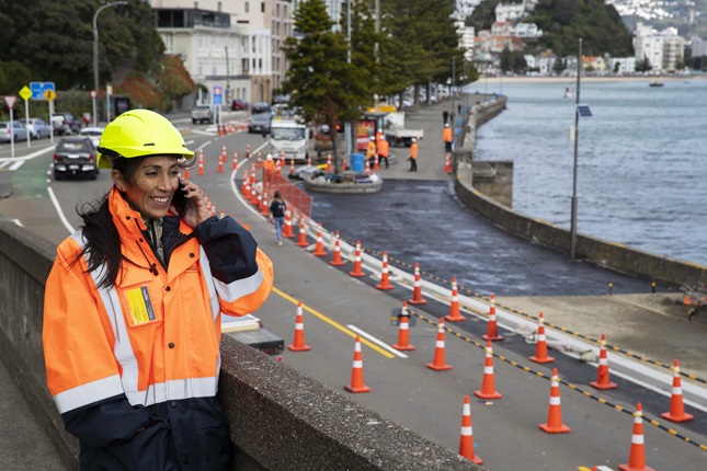 Engineer Veronica Byrne, dressed in a yellow hard hat and a hi-vis orange jacket, smiling while on the phone standing at a work site around Oriental Bay.