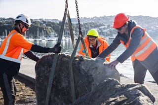 Three engineers dressed in hard hats and orange hi-vis jackets, helping position a giant boulder suspended by chain into place along the Cobham Drive.