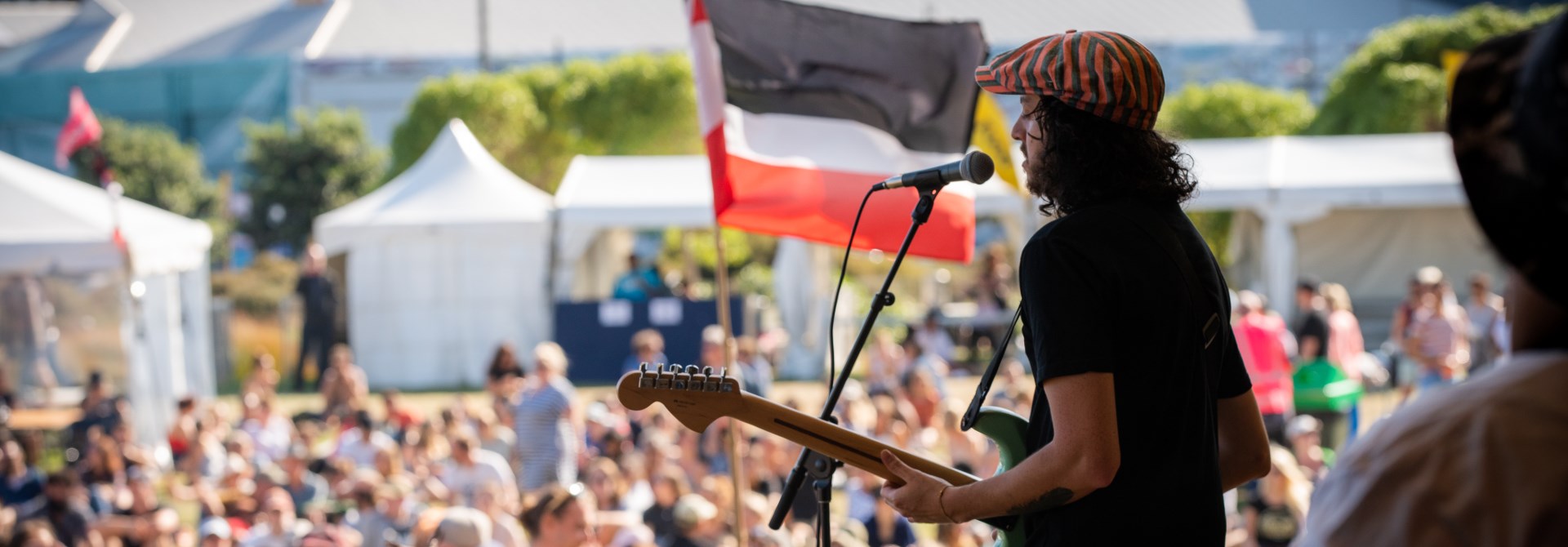 A person with a guitar signing into a microphone on stage in front of a crowd with the Māori flag waving in the background.