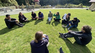Ten young people, who are members of the Zealandia Youth Collective, siting in a circle on the lawn at Zealandia.