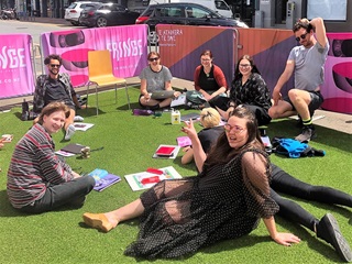 Eight adults enjoying the sunshine as they work outside on their laptops while sitting on fake green grass in the Fringe parklet, surrounded by pink fence bollards.