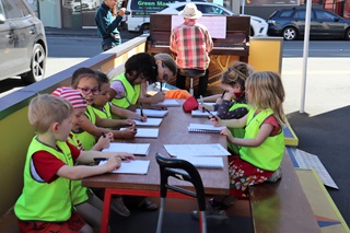 About eight young children wearing high-vis yellow vests working with paper and pens on a large wooden table, with teachers and someone playing a piano behind them.
