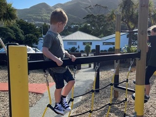 Four-year-old Jax on a yellow and black chain bridge at Karori playground, with the hills and blue sky in background.