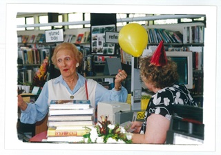 Ruth Gotlieb holding a yellow balloon in a library with another woman in a red party hat. 