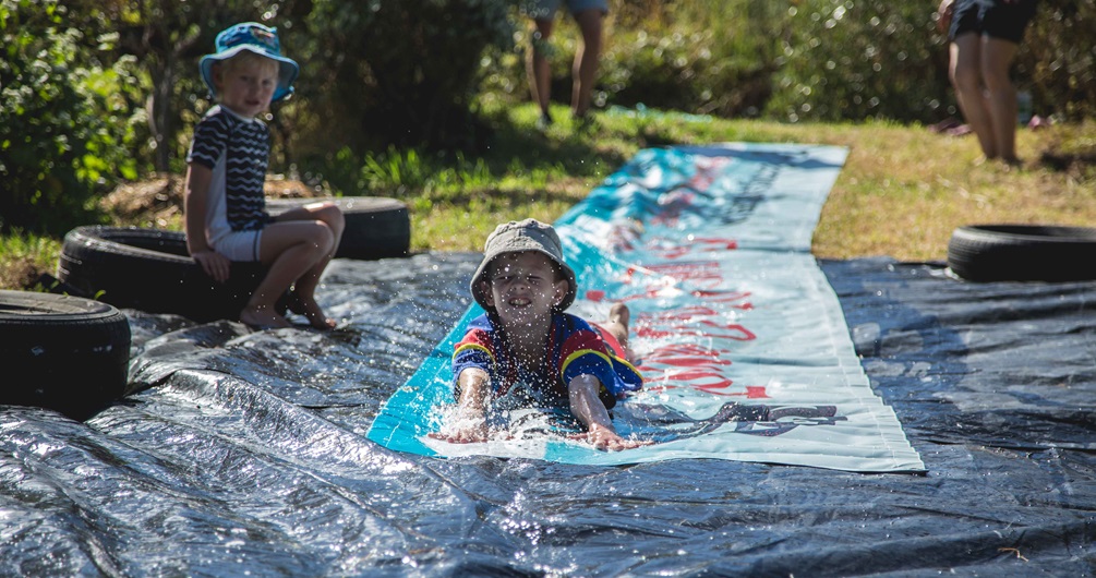 Two children in sunhats and togs, one sitting on a tyre watching on as the other slides down a waterslide tarp set up on the grass, splashing at the bottom.