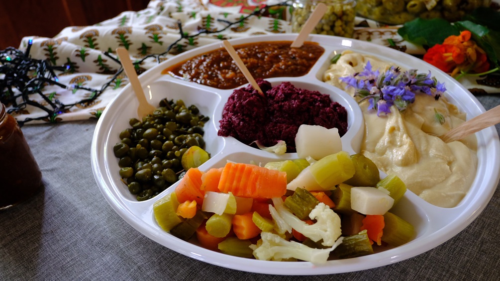 A round platter with various dips, including hummus, relish, and beetroot, with olives, vegetables, and purple flowers.