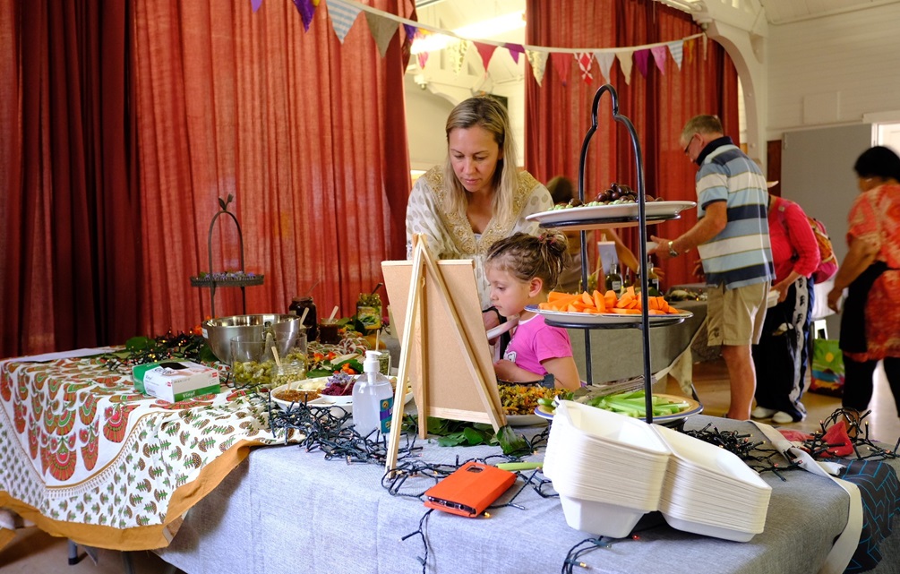 A woman and child at a table of food, with other adults serving up their plates from trestle tables in the background, in a decorated hall with large red curtains.