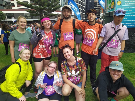 Members from Spectrum Care, all dressed in lovely bright clothing, wearing their pink registration bibs and smiling as a group while taking part in Round the Bays.