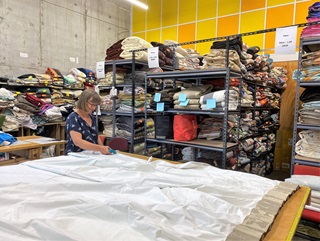 A  woman at a workbench cutting a large piece of white fabric, surrounded by shelves stacked with curtains, and yellow tiles on the wall, at the Wellington Curtain Bank.