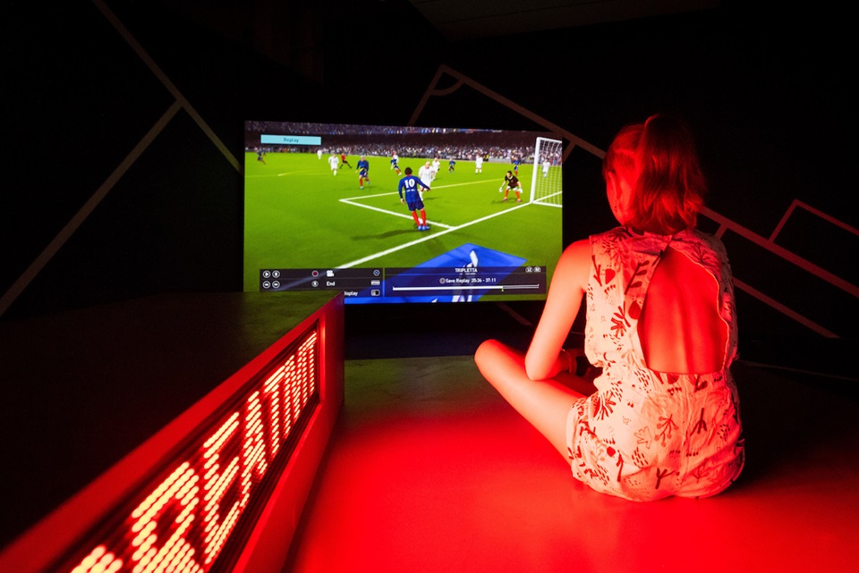 A girl sitting on a gallery floor, cast in red light from a Fluro text screen, looking at a large TV screening a football game.