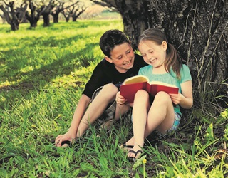 A boy and a girl smiling, huddled together reading a book under a tree on the grass, on a sunny day.