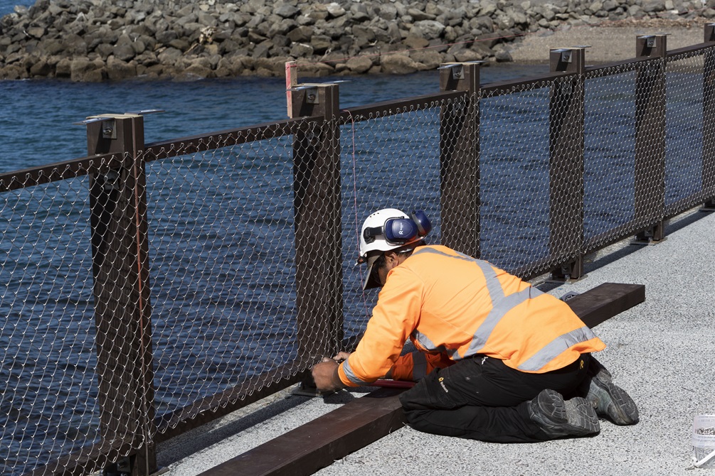 A workman kneeling on a concrete path adjusting a new fence around Kio Bay.