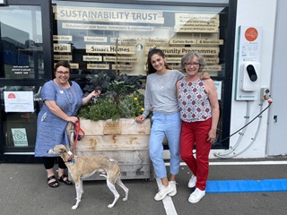 Three staff from the sustainability Trust, as well as the CEO's dog, standing in front of the small planter box vege garden, with the EV car charger on right, in front of the entrance to the trust's eco store.