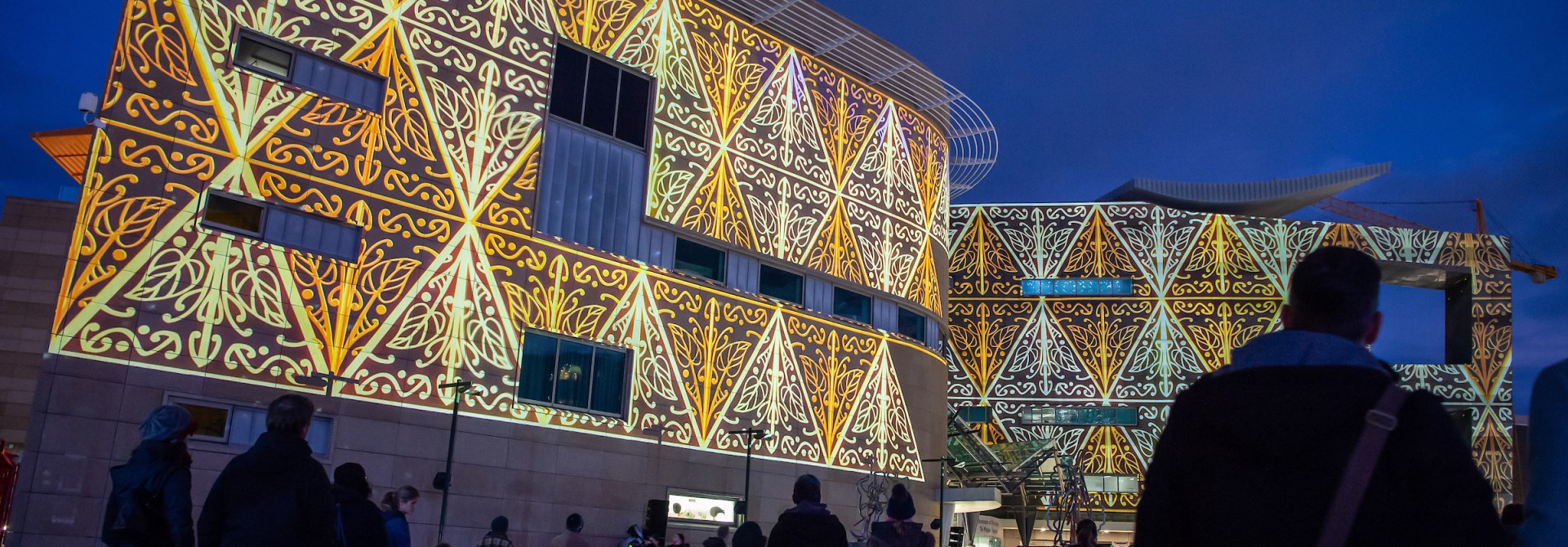 Photograph of orange and yellow Māori patterns projected onto the side of Te Papa building at night with people standing in front looking at it.