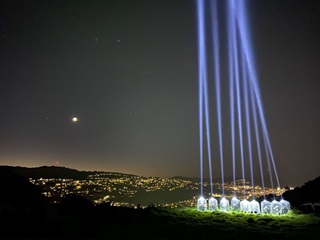 Nine Tūrama Light Beams raising to the dark night sky out of glass bottles, with the moon and Wellington's light-speckled hills in background.
