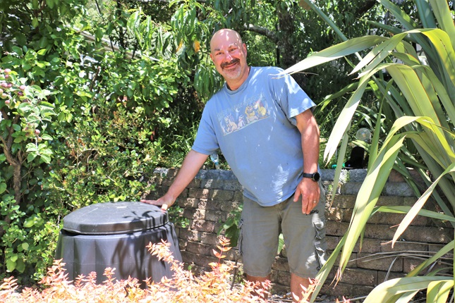 Brad from Seatoun, standing in his blue t-shirt, leaning against his black compost bin, surrounded by a tall flax bush and trees.