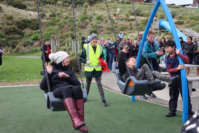 Councillor Jill Day on left, and 13-year-old Cyrus Dahl on right, swinging on swings at a park, with a crowd of people behind.