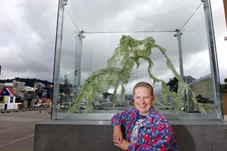 Jo Langford in a colourful red and blue jacket, leaning against a tall glass box with a green piece of 3D art inside, with Wellington waterfront in background.
