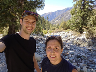 Mark Noyes and his sister, taking a selfie on rocky terrain with hilly mountains, and pine trees behind.