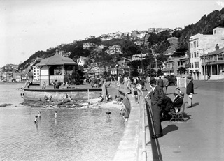 A black and white photograph of Oriental Bay and the original band rotunda in 1932, with children playing in the water below the sea wall to the left, and adults in top hats relaxing in the sun on bench seats to the right.
