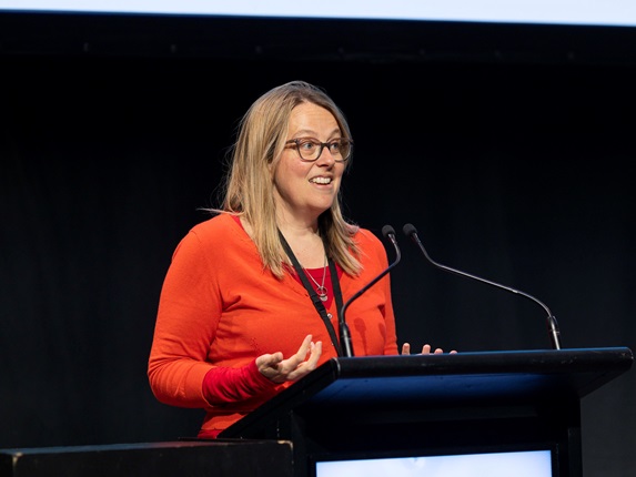 Wellington City Council's climate change response manager Alison Howard with blond hair and glasses, wearing a bright orange top and a black lanyard, looking away from the camera as she gives a talk behind a podium with black wall behind her.