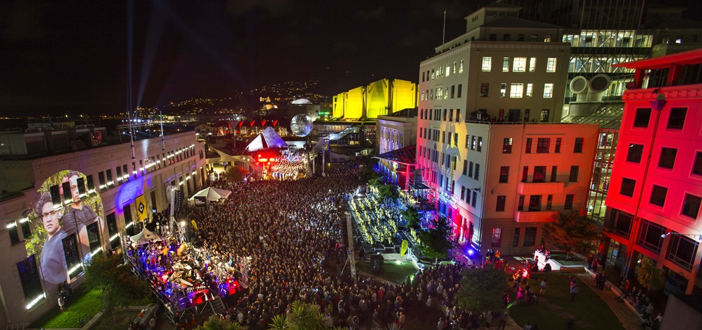 A high-angle view of a very crowded Civic Square at night time, with colourful projections on the CAB and city gallery buildings.