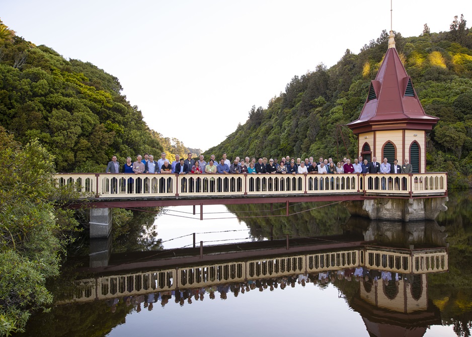 A crowd of dozens of people who have worked at Wellington City Council for 20 years or more, all standing on the cream and maroon-coloured bridge and lake lookout at Zealandia, with the structure and people reflected in the water below. Picture taken in early 2000s.