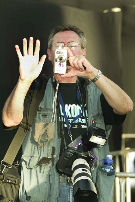 Photographer Neil Price holding his left palm up while the other hand holds a small digital camera, as he concentrates on the frame he is capturing. Neil has a green vest on with a drink bottle stuffed in the pocket and a larger canon DSLR camera around his neck. 