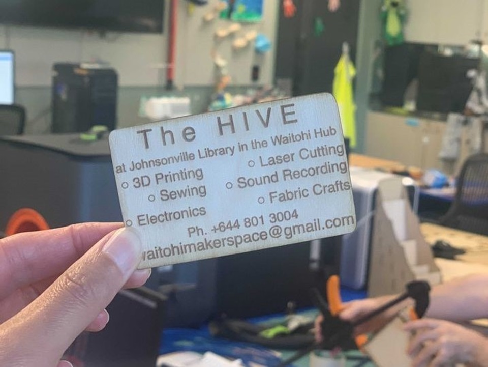 A hand holding up a business card for The Hive at Johnsonville Library in the Waitohi Hub, detailing the services it offers which includes 3D printing, sewing, laser cutting, and sound recording, with the working space in behind the card. 