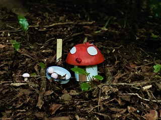 A hand made little red mushroom nestled into the leaves on the ground with a dish beside it containing three small eggs.
