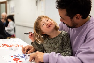 A father and young daughter sitting at a table doing art together, smiling into each others faces.