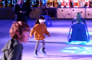 Young children wearing warm clothes and skating on an indoor ice rink with bright lights all around.