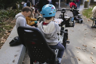 The back of a young boy with a grey woolly hoodie and a bright blue helmet, sitting in a black child's seat on the back of an adult's bike with a clear lunchbox, and adults sitting on a concrete bench to his left.