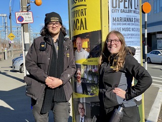 Two Wellington parking officers, Jacob and Caitlin, in their black uniforms, smiling and leaning against a billboard-plastered pole in the middle of a street with bright blue sky, parked cars, and a pedestrian crossing behind.