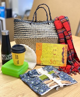 A photograph of a collection of items on a table including a whicker basket, a red tartan scarf, a black waterbottle, a yellow reusable cup, a green tupperware container, a $50 Tip Shop voucher, a material food bag and a bamboo cutlery set.
