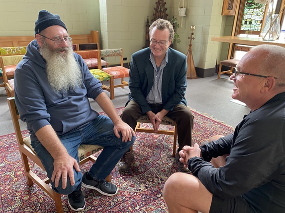Paul sitting with a long grey beard and beanie on his head, with Alex on his right seated on a chair, neatly dressed and smiling, and Ray Tuffin with short hair and glasses, also seated, facing the other two having a chat on a large red Persian rug and church pews behind.