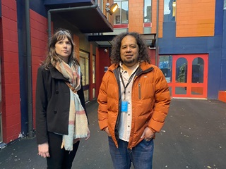 A woman in a clack coat and colourful scarf standing next to a man with long black curly hair and an orange jacket and blue jeans, in front of a bright blue, red and orange building.