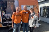 Two older gentlemen in orange t-shirts and a woman in a grey hoodie - all tops have the words Orange Sky Aotearoa on them - smiling with their arms around one another, with an orange van behind them.