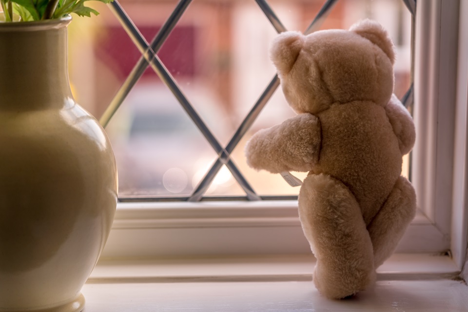 A small light brown teddy bear looking out of a window, next to to a vase of flowers.