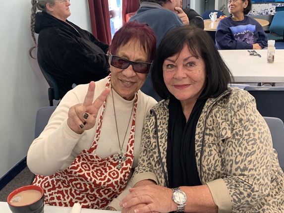Two women sitting beside each other, Teia on the left wearing her sunglasses and an apron doing the peace sign, and Amber leaning in smiling.