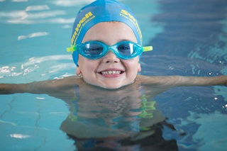 A young child wearing a blue swim cap and goggles, floating in a pool and smiling.