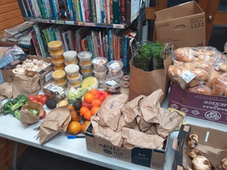 A table filled with donated food, including soups, fresh produce, breads and containers of olives and meat, in front of a library shelf filled with books.
