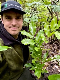 A selfie of a young man who looks to be in his 30s, wearing a black cap and dark green jacket, in a bush setting nestled in between branches and leaves of a kohekohe tree.