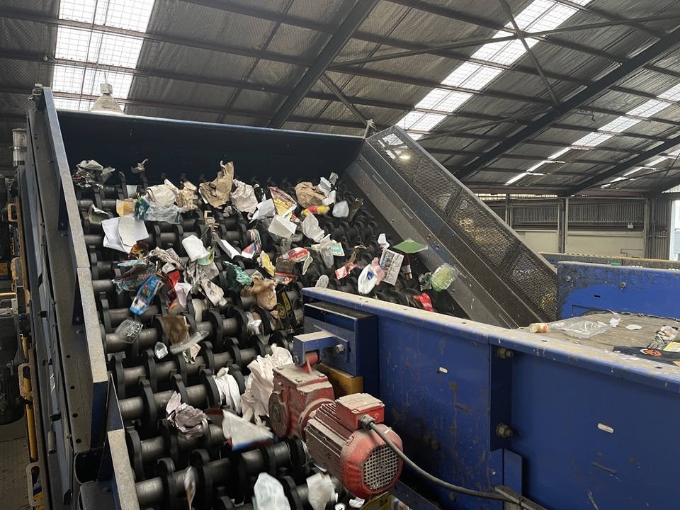 A large steeply-angled tray filled with recycling bottles, containers and other recyclable materials, feeding onto a blue metal conveyor belt, in a large warehouse with clear light panels on the roof.