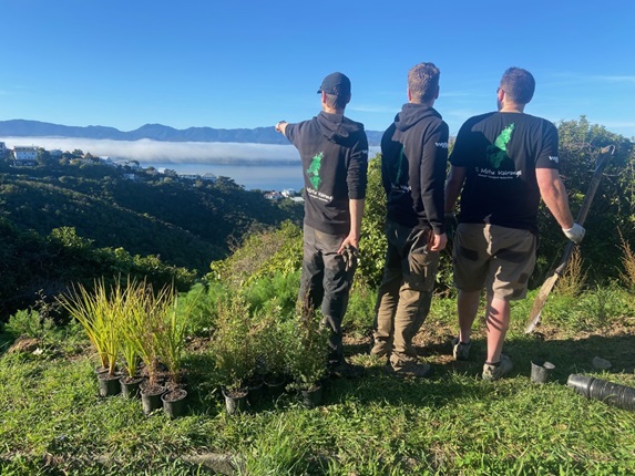 Three men on a grassy hill, wearing matching black hoodies with restoration group Te Motu Kairangi braded on the backs of them, overlooking Wellington hills masked in low-lying cloud on a blue-sky day.