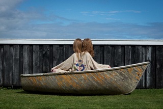 An artowrk of two girls sitting back to back in a small woodnen boat on a green lawn, both wearing a kimono with a dragon on it.