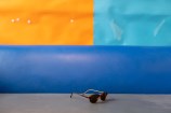 A pair of brown sunglasses abandoned on a blue and grey couch with colourful squares hanging on the wall in the background.