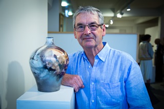 A man wearing glasses and a blue shirt leans on a plinth next to a marbled, glazed vaze in a gallery.