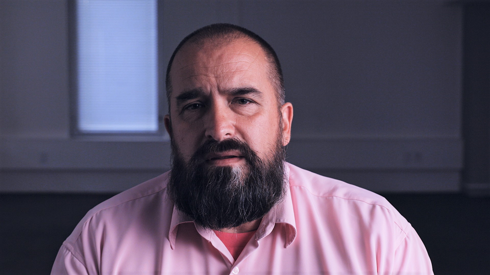 A man with a dark beard and pink shirt sitting in a dark room and talking direct to camera.