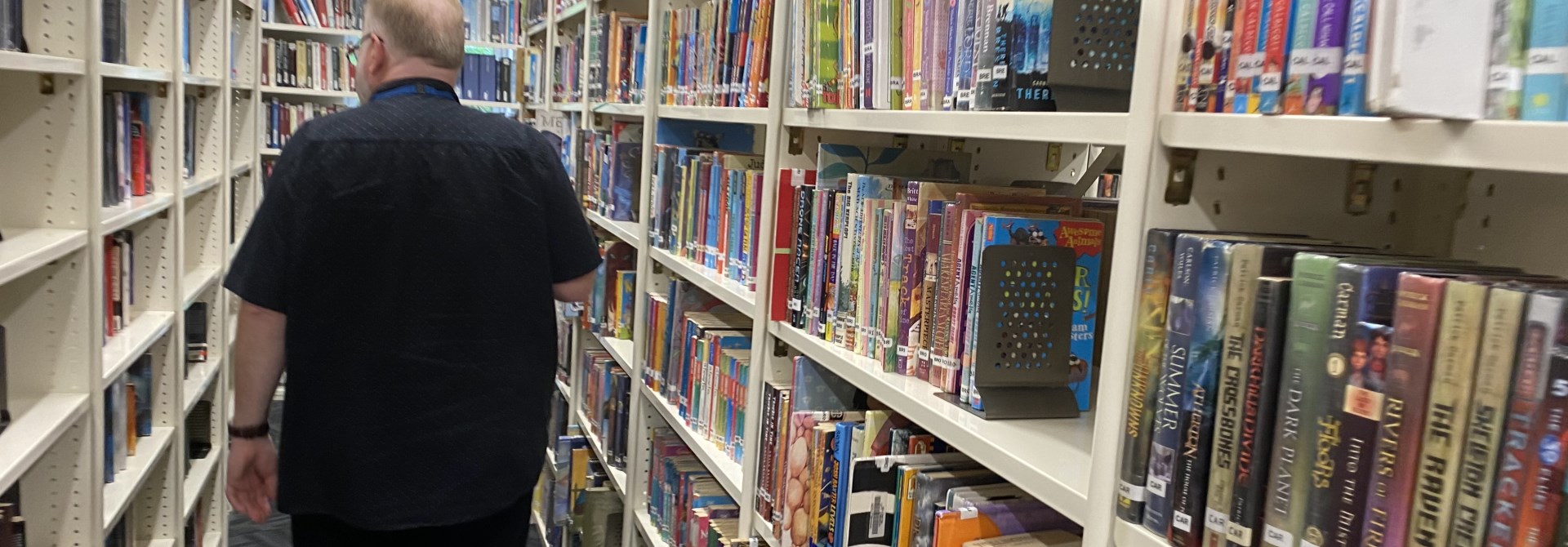 The back of a large-framed man with dark coloured clothing walking through a library isle of shelving filled with books on either side.